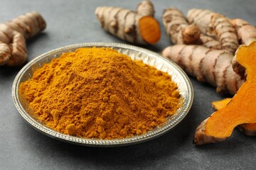 Plate with turmeric powder and raw roots on grey table, closeup