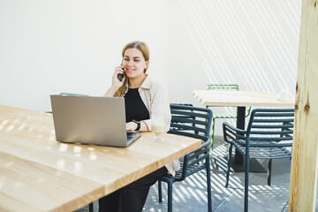 Young happy woman sitting at outdoor cafe table and talking on phone with cup of coffee, smiling woman enjoying telecommuting in cafe or studying online on laptop