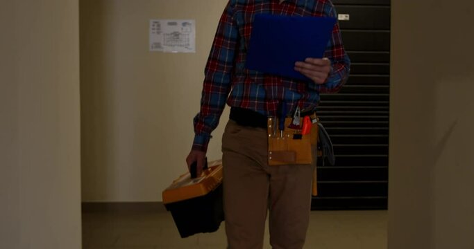 Male repair worker walks down corridor after completing order. Man employee in work uniform holds clipboard and toolbox in apartment complex