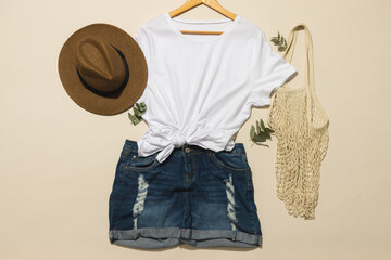 Flat lay of white t shirt, sunhat, net bag and denim shorts with copy space on cream background