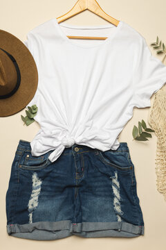 Flat lay of white t shirt, sunhat, net bag and denim shorts with copy space on cream background