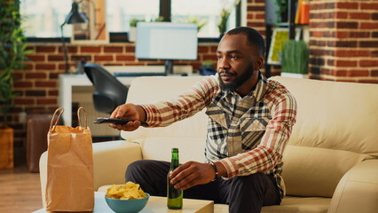 Relaxed guy having meal from fast food takeout place, unpacking food from delivery bag in front of television. Young person preparing to eat dinner and watch comedy tv show or movie on sofa.