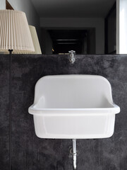 Empty big white vintage medical wall mount bathroom sink on black concrete wall decoration near big mirror and standing light lamp in the bathroom, vertical style. White farmhouse sink bathtub design.