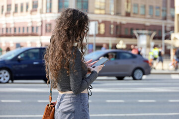 Girl with long curly hair standing with smartphone on a street on cars background. Mobile communication in city