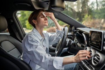 Fototapeta na wymiar Woman drives car with broken air conditioning in hot summer weather wipes sweat on forehead with paper napkin. Middle aged female suffering from heat stuffiness. Exhausted overheated stressed driver.