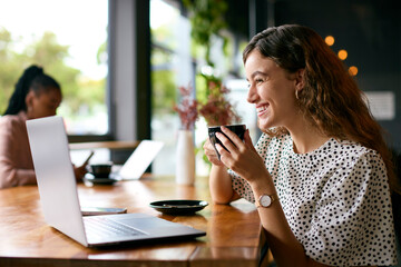 Young Businesswoman With Coffee Working On Laptop Sitting In Cafe With Colleague In Background