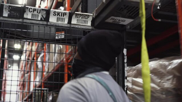 A BIPOC warehouse distribution center merchandise worker drives an industrial forklift picker down a racked DC isle to retrieve furniture inventory to be shipped.