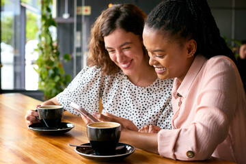Two Young Female Friends Meeting In Coffee Shop And Looking At Mobile Phone Together
