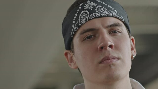 Medium closeup portrait of confident young Caucasian male rapper in black and white bandana hat and earring in his ear looking at camera confidently outdoors in street
