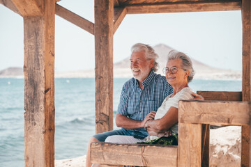 Lovely senior family couple sitting close to the beach looking at horizon over sea, two elderly people enjoying vacation or retirement together
