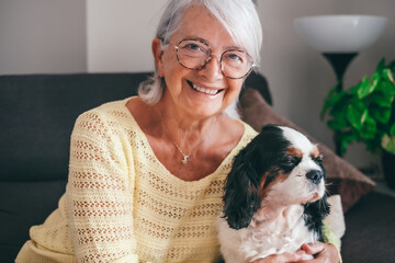Smiling senior woman sitting on sofa at home while hugging her cavalier king charles spaniel dog. Elderly retired lady with her best friend looks into the camera