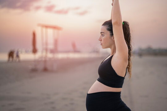 Side view portrait of pregnant woman in black sportswear making Crescent Lunge yoga pose on the beach at sunset. Working out, yoga and pregnancy concept.