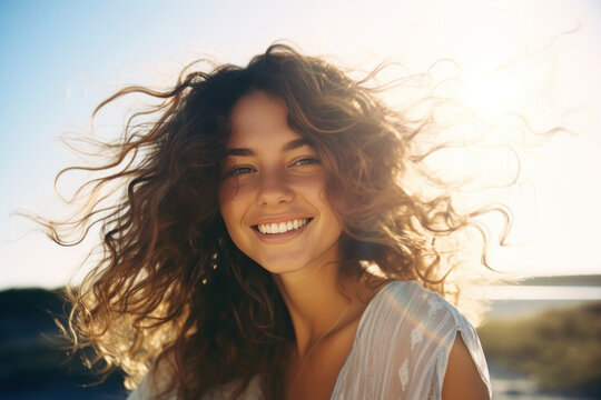 jeune femme souriante avec des cheveux bruns et bouclés dans le vent, prise de vue à contre-jour une journée d'été ensoleillée, ciel bleu