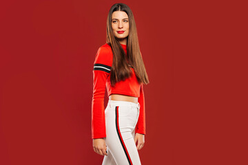 Fototapeta premium Attractive trendy young hipster with long brunette hair, in white trousers and red shirt, looking away on red background.