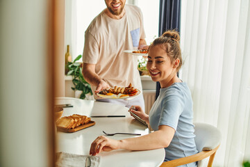 Smiling woman sitting near cutlery and blurred boyfriend holding tasty breakfast at home in morning
