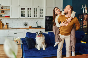 Smiling woman piggybacking on boyfriend near border collie dog on couch and popcorn in living room