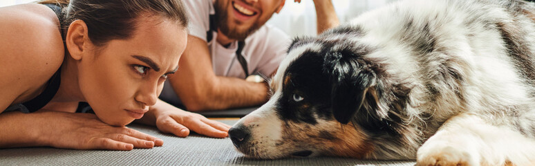 Displeased woman looking at border collie near smiling boyfriend on fitness mat at home, banner
