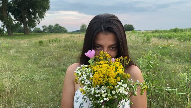 Portrait adorable young woman enjoys leisure time in nature beauty at picturesque meadow, inhales odorous smell of wildflowers. Cheerful female person in dress looking at camera, smiling charmingly