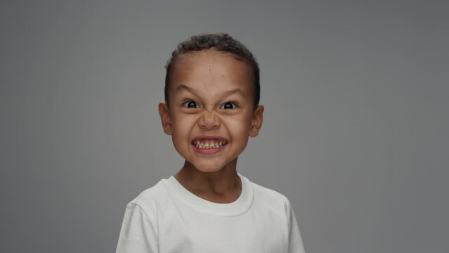 Portrait of Young Boy Looking at Camera with Angry Grin in Color Studio Shot. African American Kid or Mixed Race Child Isolated Alone on Grey Background Close-up. Male Person with Upset Face Grimace