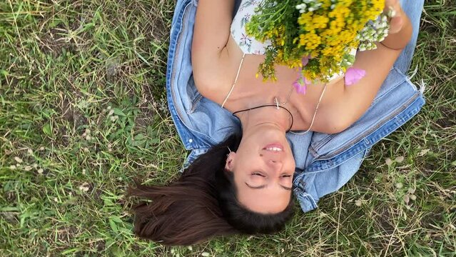 Beautiful young woman enjoys effortless pastime in nature beauty. She lying on back on grass meadow, holds colorful bouquet of wildflowers, smells odorous wild herbs. Directly above rotating shot
