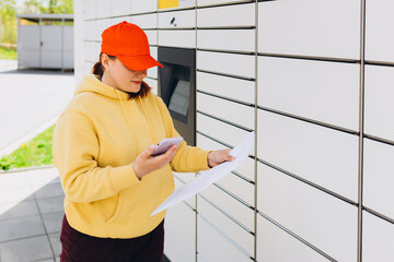 Young woman in red hat picks up parcel from automatic post office machine, Courier standing with phone and small box. Concept of fast delivery to automatic self lockers
