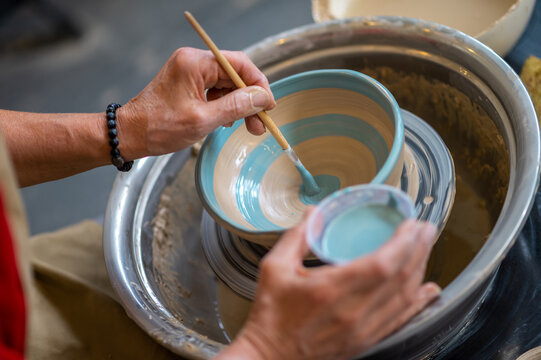 Closeup of woman painting clay mug, coloring pottery in workshop with a paintbrush.