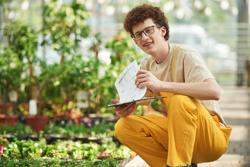Quality control, with notepad. Young man with curly hair and in glasses is in greenhouse