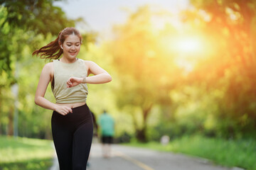 Beautiful Asian female jogger running in the park and looking at her watch to see her running timing in the park background.