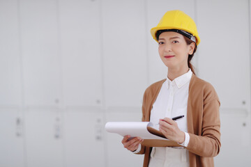 Beautiful young woman engineer wearing white shirt brown blouse and yellow safety helmet holds a pen with clipboard.