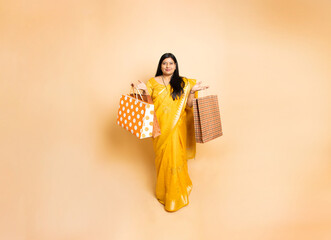 Happy young beautiful indian woman wearing sari holding shopping bags standing isolated on beige...