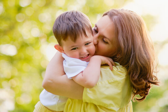 A mother woman kisses a baby boy on the cheek while walking in nature. Toddler baby and mom play on the green grass in the summer park. Kid aged about two years (one year eleven months)