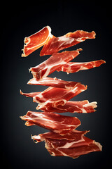 Thin slices of Spanish ham jamon flying in the air on black background. Traditional meat specialty of the local cuisine - obrazy, fototapety, plakaty