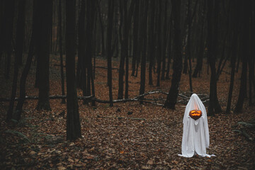 Boo! Spooky ghost holding glowing jack o lantern in moody dark autumn forest. Person dressed in...