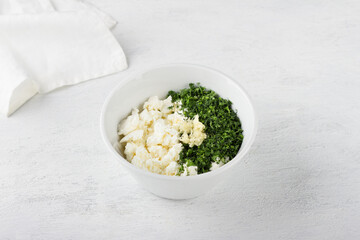 White bowl with cottage cheese cream mixed with herbs and garlic on a light gray background. Cooking appetizer or toppings for stuffing vegetables, vegetarian food
