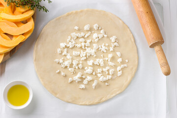 Rolled out dough with crumbled feta cheese, bowl with olive oil, rolling pin, cutting board with sliced pumpkin and fresh thyme on white background. Cooking step of delicious homemade pumpkin pie - 630332419