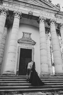 Newlyweds walk near Baroque Roman Catholic church in Pidhirtsi Ukraine. Bride and groom kissing on stairs near large columns of ancient temple at sunset. Couple kiss. Back view. Black and white photo