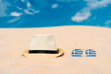 Sunglasses with glasses in the form of the flag of Greece and a hat lie on the sand against the blue sky. The concept of summer holidays, travel and tourism in Greece