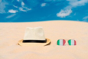 Sunglasses with glasses in the form of the flag of Italy and a hat lie on the sand against the blue sky. The concept of summer holidays, travel and tourism in Italy