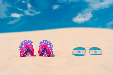 Sunglasses with glasses in the form of the flag of Argentina and flip-flops lie on the sand against the blue sky. The concept of summer holidays, travel and tourism in Argentina