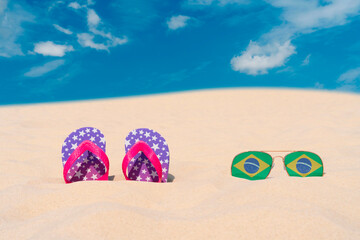 Sunglasses with glasses in the form of the flag of Brazil and flip-flops lie on the sand against...