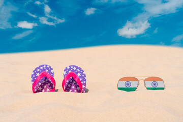 Sunglasses with glasses in the form of the flag of India and flip-flops lie on the sand against the blue sky. The concept of summer holidays, travel and tourism in India