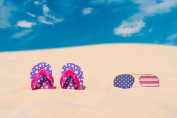 Sunglasses with glasses in the form of the flag of USA and flip-flops lie on the sand against the...