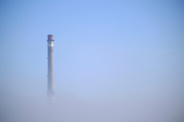 Thick fog from which a factory with a large industrial pipe is visible