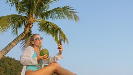 Natural beauty woman spends her holidays on tropical island near palm tree.Woman use mobile phone, take photo self for social media.Woman make selfie.Tourist enjoy vacation and drink coconut cocktail