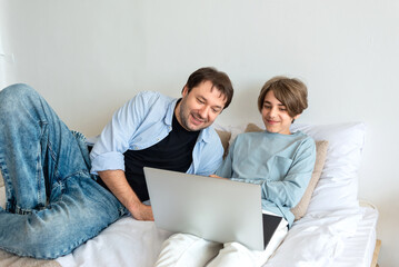Father and teenager son looking at laptop screen, smile, watching video, communicate online together, in social media, laying on bad at home. Happy family relations, child support, fatherhood concept