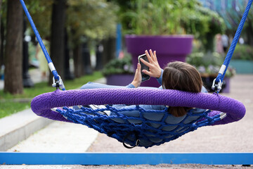 Girl lying with smartphone in swing, leisure in summer park