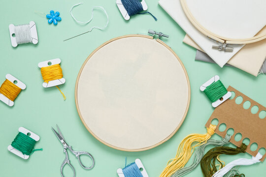 Embroidery set fot stitching. Beige cotton cloth in embroidery hoop on green background with fabric, colorful threads, scissors and needls. Indoor hobby concept with copy space, top view