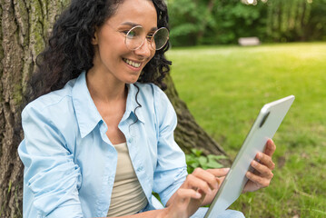 Portrait of a young Middle Eastern woman in casual wear wearing glasses who uses a tablet to touch and read good news. A girl in the park sits on the grass enjoying a wonderful holiday and the sun