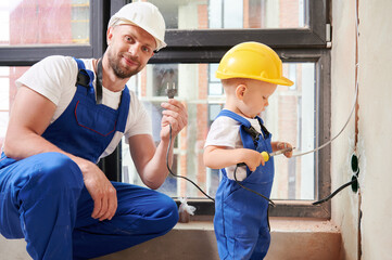 Male electrician holding electrical plug and smiling while child using electric soldering iron tool. Man and kid wearing work overalls and safety helmets while working on home renovation. - Powered by Adobe