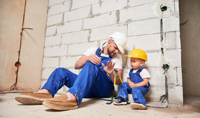 Male builder in work overalls showing pliers to little boy in apartment under renovation. Man and...